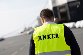 Ein Mann, der mit dem Rücken zur Kamera steht, trägt eine gelbe Warnweste mit der Aufschrift ,,Anker“.