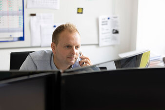 Ein Mitarbeiter sitzt vor dem PC und hält ein Telefon am Ohr.